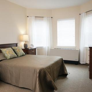 A cozy bedroom with a neatly made bed, bedside lamp, and ample natural light from the windows.