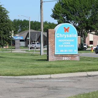 A sign for Chrysalis placed on a grassy area near a road, with buildings in the background.