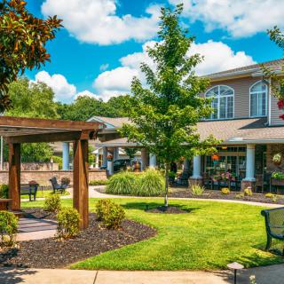 A beautifully landscaped garden with a wooden pergola and outdoor seating adjacent to a building under a bright blue sky.