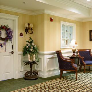 A warmly decorated hallway with a seating area, featuring chairs, a small table with a lamp, and decorative elements including a wreath on the door.
