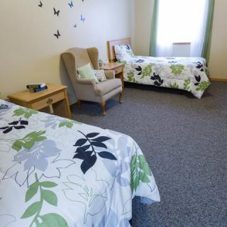 A cozy bedroom with twin beds featuring floral bedding, an armchair, and a window with curtains.