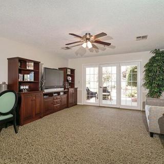 A cozy living room with a ceiling fan, large bookshelf, television, and ample natural light from large windows.