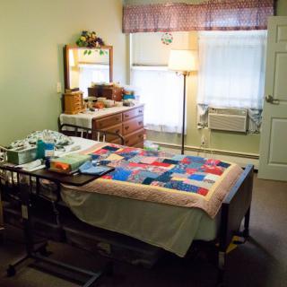A cozy bedroom with a quilt-covered bed, dresser, and soft lighting from a floor lamp.