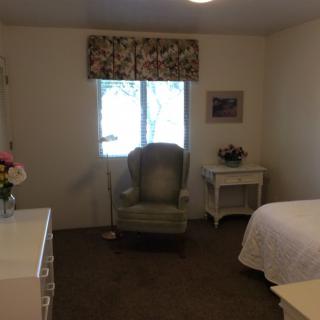 A cozy bedroom featuring a green armchair by the window, a bedside table with flowers, and a neatly made bed.