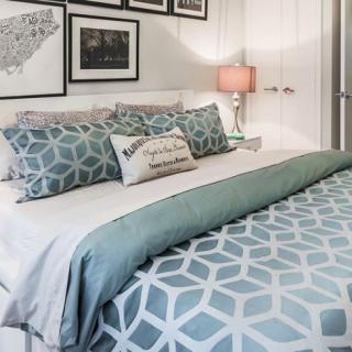 A cozy bedroom with geometric-patterned bedding and decorative pillows, complemented by framed wall art and a soft-lit lampshade.