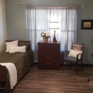 A cozy bedroom featuring a single bed, dresser, and a comfortable chair with natural light streaming through the curtains.
