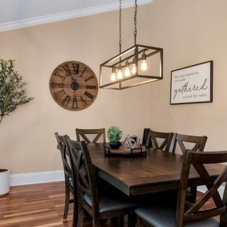 A cozy dining room with a wooden table and chairs, featuring a pendant light and decorative elements like a wall clock and a potted plant.