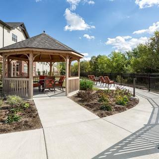 A scenic outdoor area featuring a gazebo with seating, surrounded by a landscaped garden and pathway under a clear blue sky.