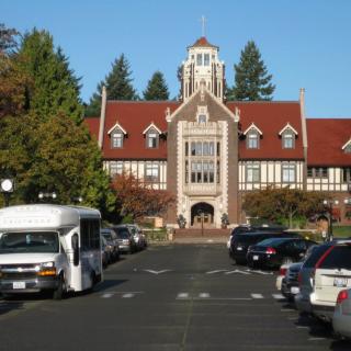 A historic building with a red roof and tower, viewed from a parking lot bordered by trees.