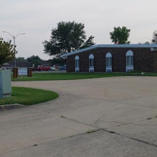 A view of a brick building with arched windows, surrounded by greenery and a paved road.