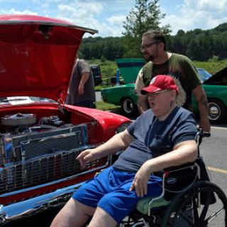 A man in a wheelchair enjoys the sights at a local car show, with a vintage red car on display.