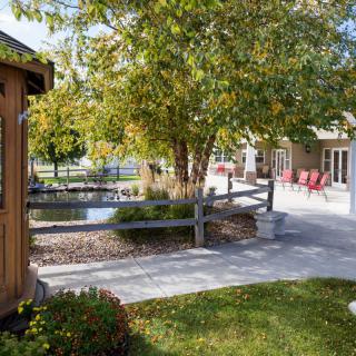 A serene outdoor garden with a pathway, surrounded by trees and a wooden fence, leading to a seating area.