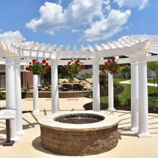 A beautifully designed outdoor space featuring a stone fire pit surrounded by a classic white pergola and lush greenery under a bright blue sky.