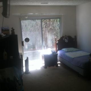 A dimly lit bedroom with a single bed and sunlight streaming through a large window.