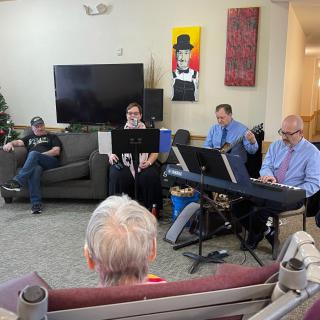 A small band performs in a cozy living room setting for an audience.