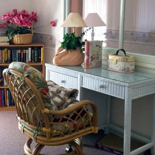 A cozy home office setup features a wicker desk and chair, with a lamp, books, and a basket of flowers creating a welcoming atmosphere.