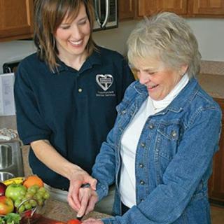 A caregiver helps an elderly woman prepare food in a warm kitchen setting.
