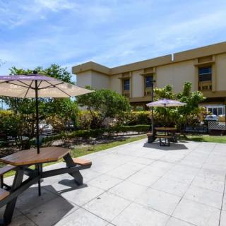 A sunny outdoor patio area with picnic tables, umbrellas, and lush landscaping in front of a building.