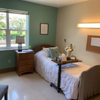 A cozy and well-lit bedroom featuring a bed with a bedside table, lamp, and a view outside.