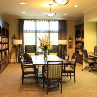 A cozy and organized office space featuring a large wooden table surrounded by chairs, complemented by bookshelves and ample natural light.
