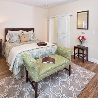 A cozy bedroom with a neatly made bed, decorative pillows, a cushioned bench at the foot of the bed, and a small table with a vase of flowers.
