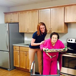A caregiver assists an elderly woman in the kitchen as they prepare to bake.
