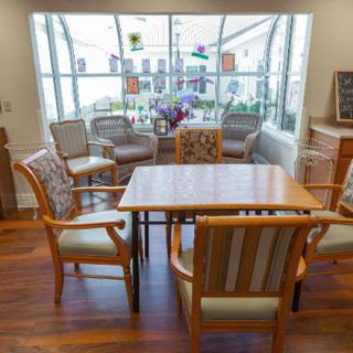 A cozy living room setup with a wooden table surrounded by chairs, decorated with flowers and colorful window ornaments.
