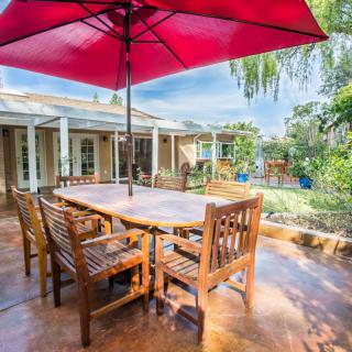 A serene backyard patio featuring a wooden dining set under a vibrant red umbrella, surrounded by lush greenery.