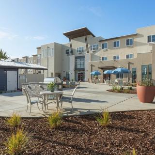A modern building with an outdoor patio area featuring tables, chairs, and landscaping.