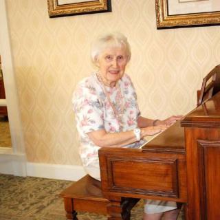An elderly woman sitting and smiling while playing the piano in an indoor setting.