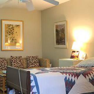 A cozy bedroom featuring a bed with a colorful quilt, a nightstand with a lamp, and wall artwork.