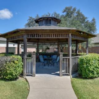 A wooden gazebo surrounded by greenery with outdoor seating inside, set under a clear blue sky.