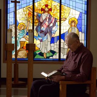 An elderly man engages in quiet reflection while reading in front of a beautifully crafted stained glass window.