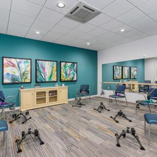 A modern exercise room with chairs arranged around hand weights, artwork on a teal accent wall, and a large mirror.