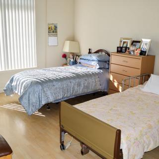 A bedroom featuring two beds, one resembling a hospital bed, with a dresser and window blinds.