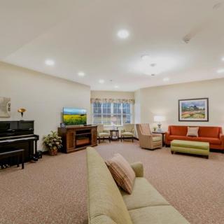 A spacious and well-lit living room featuring a piano, television, multiple sofas, armchairs, and a large window.
