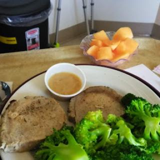 A plate of sliced cooked meat served with broccoli and a small bowl of dipping sauce, accompanied by a side of fresh cantaloupe.