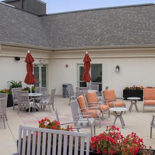 A rooftop patio featuring orange-cushioned chairs, tables with red umbrellas, and vibrant flower arrangements.