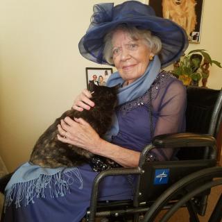 An elderly woman in a wheelchair, wearing a blue hat and dress, holding a cat.