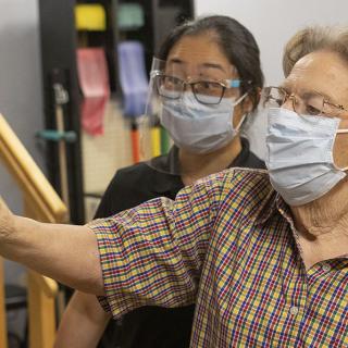 A healthcare professional assists an elderly person during a physical therapy session, both wearing masks for safety.