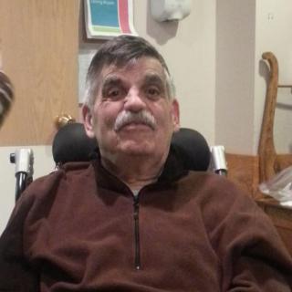 An older man with a mustache sitting in a wheelchair indoors.