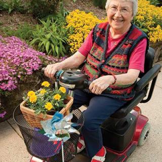 An elderly woman enjoying a ride on her mobility scooter, surrounded by blooming flowers.