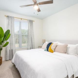 A bright and cozy bedroom featuring a large bed with decorative pillows, a ceiling fan, and a window with sheer curtains allowing natural light to enter.