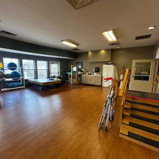 A spacious activity room equipped for rehabilitation and exercise, featuring wooden flooring and various equipment.