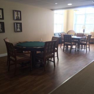 A dining area with wooden floors, featuring several tables and chairs, illuminated by natural light from large windows.