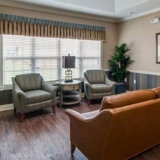 A cozy living room with a sofa, two chairs, a side table with a lamp, and large windows with curtains.