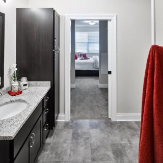 A modern bathroom with a granite countertop, dark cabinets, and a red towel. The room opens into a well-lit bedroom.