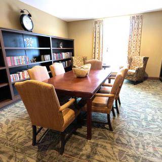 A cozy library room with bookshelves, a large table surrounded by chairs, and a carpeted floor.
