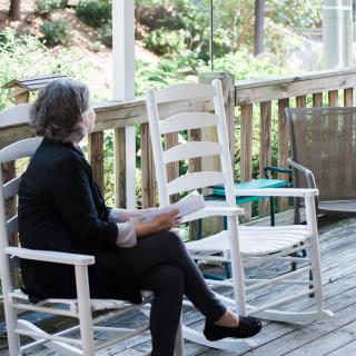 A woman sits on a wooden deck in a rocking chair, reading and enjoying the outdoors.