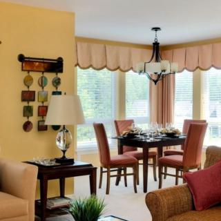 A cozy living room with an adjacent dining area, featuring a table set for four and decorated with warm tones and natural light from large windows.